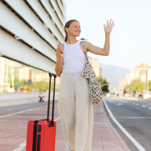 Young Beautiful Woman With Red Suitcase With Smartphone Raising Hand To Catch Taxi Cab At Terminal Or Station.