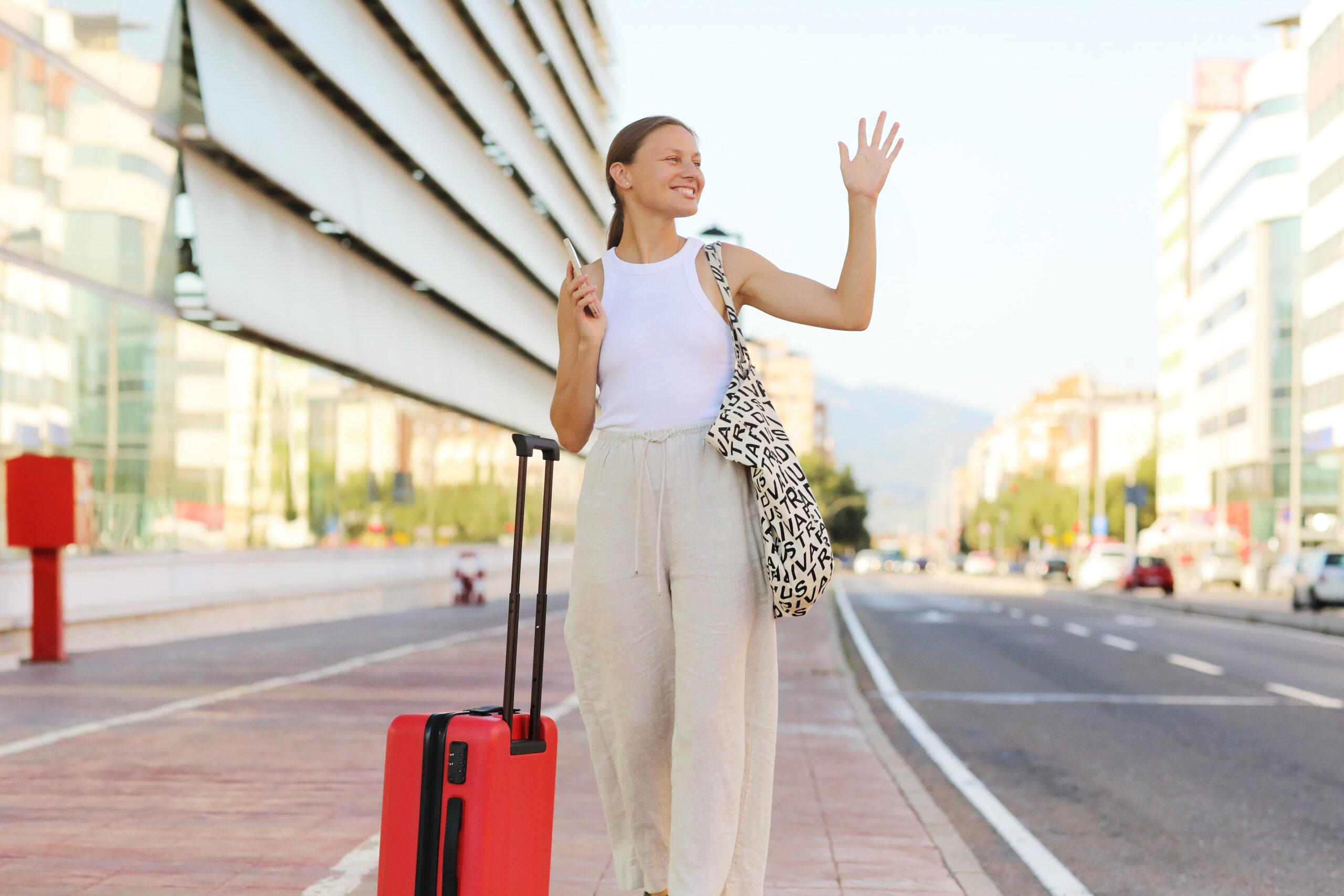 Young Beautiful Woman With Red Suitcase With Smartphone Raising Hand To Catch Taxi Cab At Terminal Or Station. Stylish Smiling Girl Tourist On Summer Holiday Vacation. New Trip